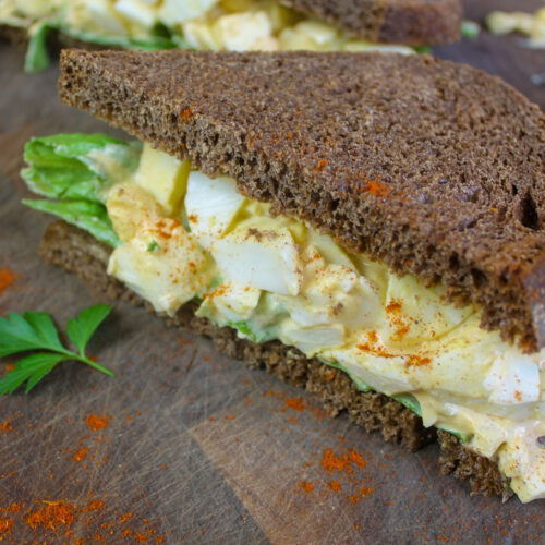 Egg salad sandwich on dark pumpernickel bread on a wooden cutting board.
