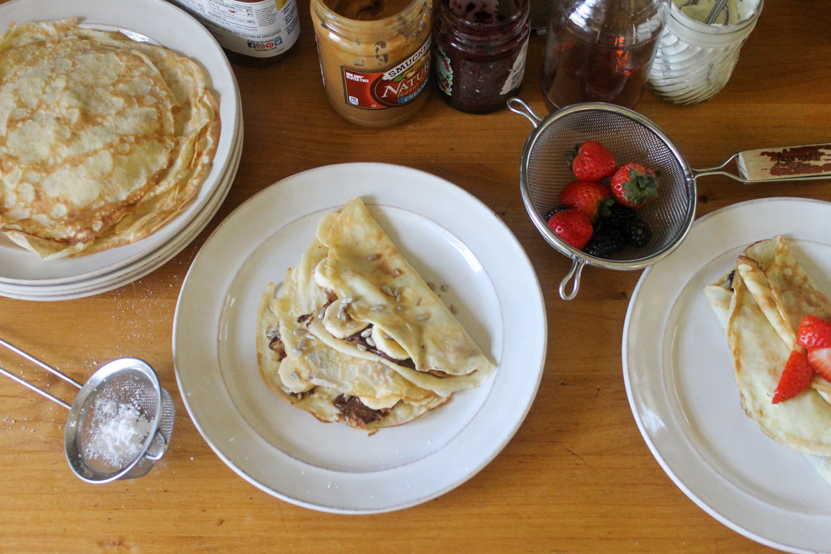 Crepes being assembled on the counter top, with topping choices like berries, peanut butter, syrup, Nutella and powdered sugar.