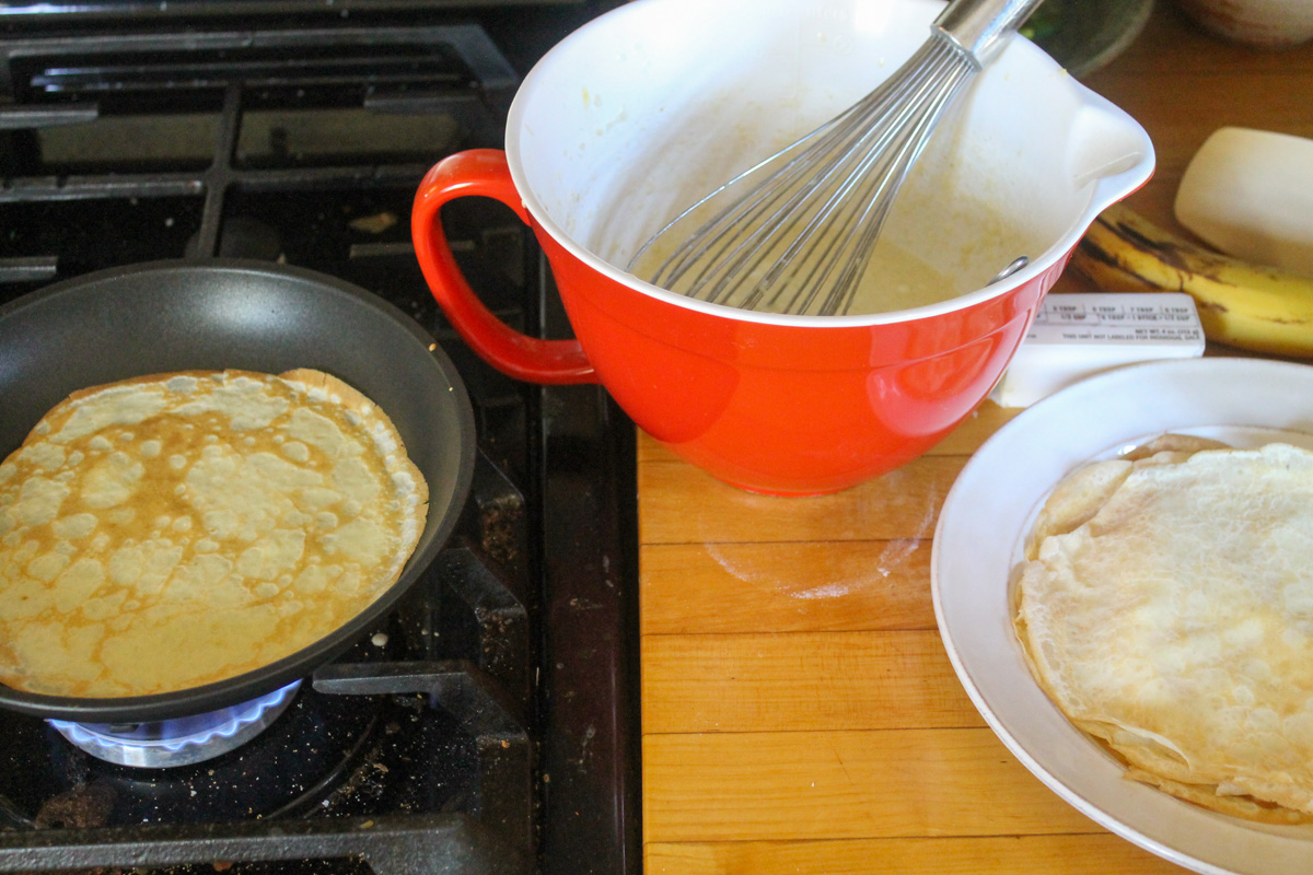 A crepe cooking in a non-stick skillet on the burner next to a red bowl of batter and a plate of finished crepes.