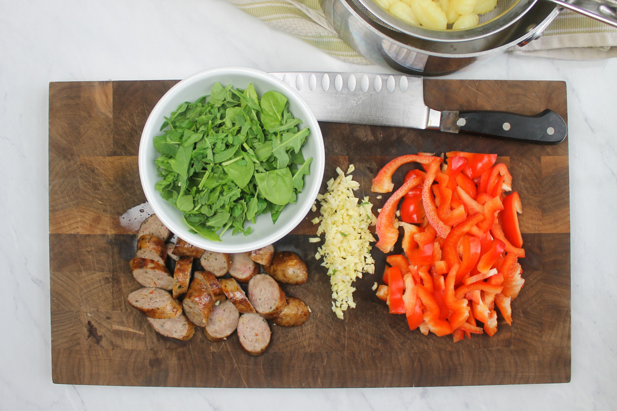 Ingredients chopped on a cutting board, sliced red bell pepper, minced garlic, sliced sausages, and roughly chopped greens.