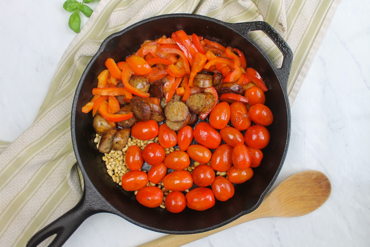 Cherry tomatoes and pine nuts added to the red bell pepper and sausage in a black skillet.