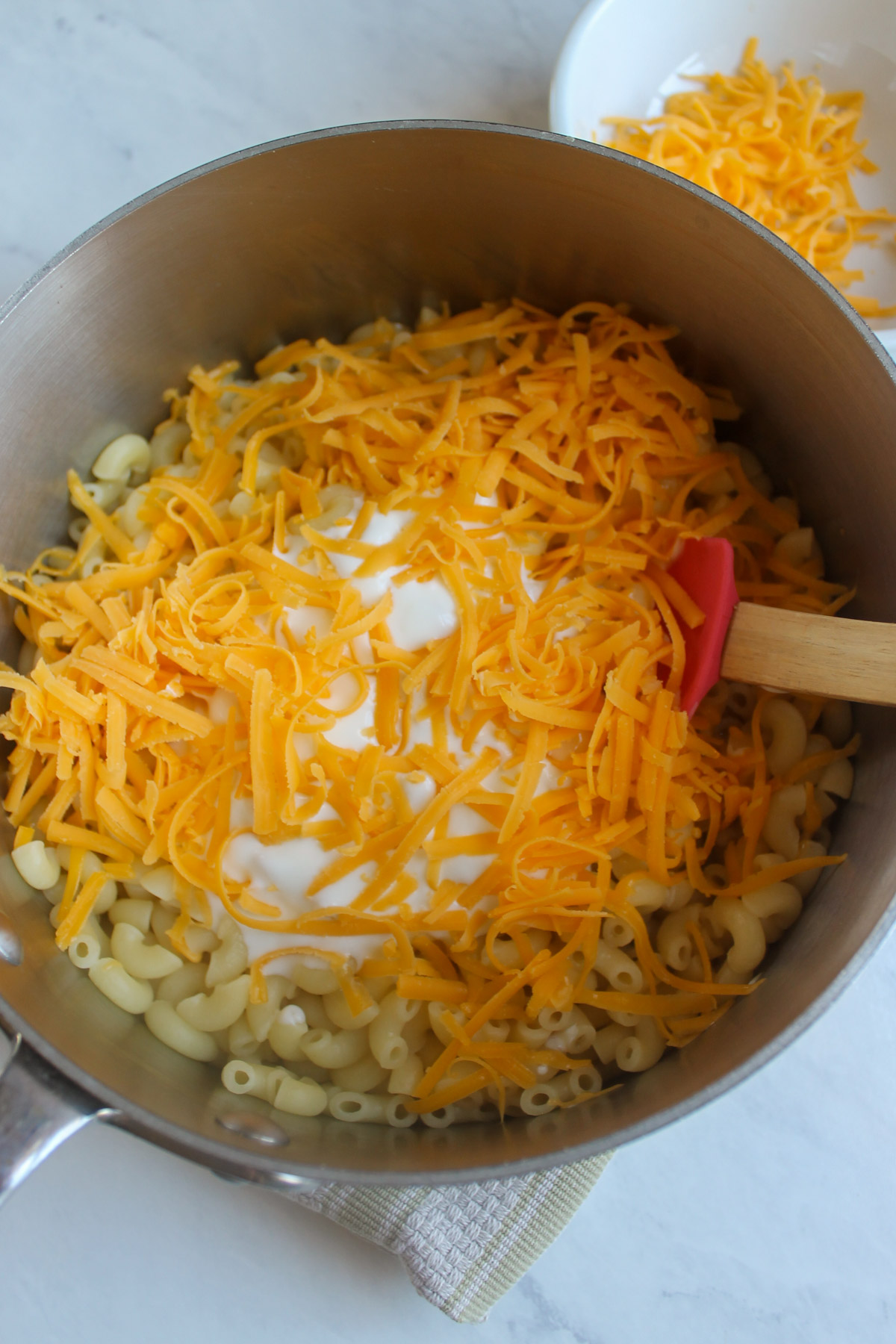 Adding yogurt and shredded cheddar cheese to a pot of boiled macaroni pasta.