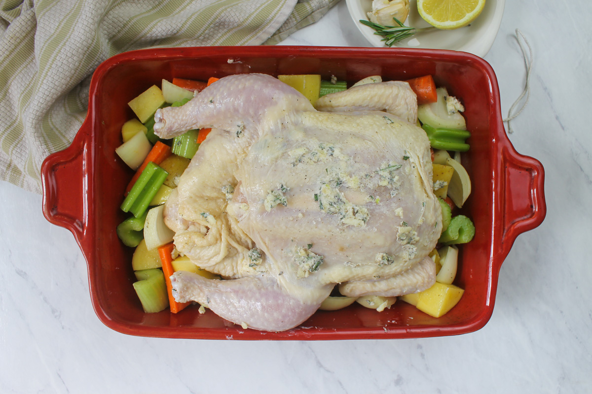 A raw whole chicken smeared with garlic butter on a bed of raw vegetables.