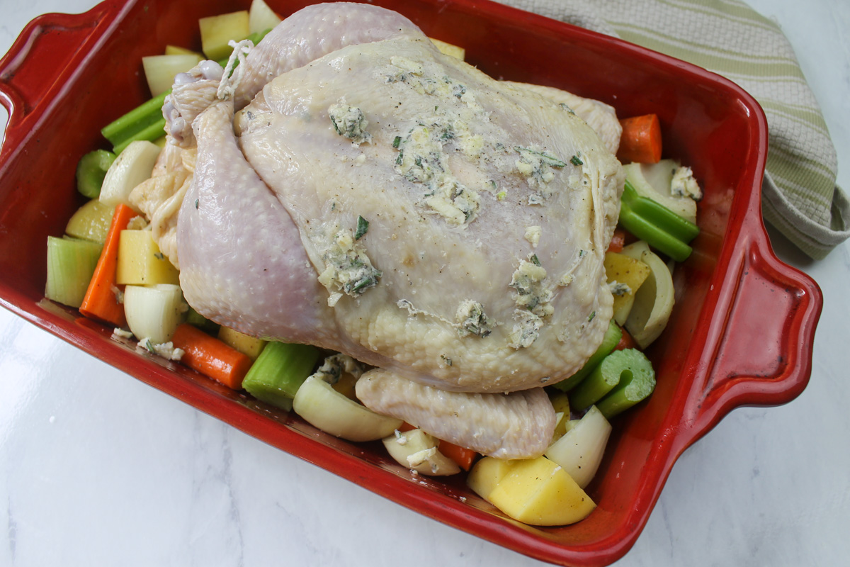 A whole raw chicken with legs tied, on a bed of root vegetables ready to roast.