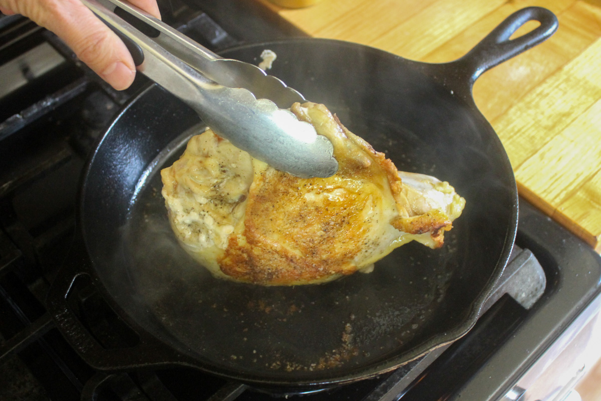 Tongs holding up a chicken breast to sear all sides in a cast iron skillet.