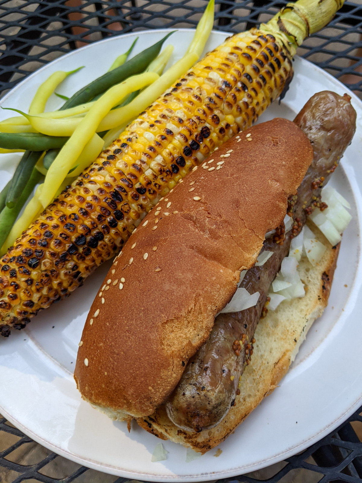 A dinner plate with charred corn on the cob, a brat in a bun and garden green beans.