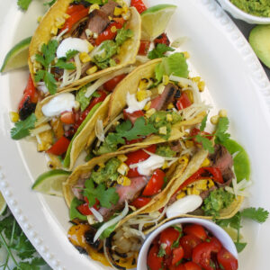 A white platter of steak fajita tacos with charred corn, and grilled peppers and onions.