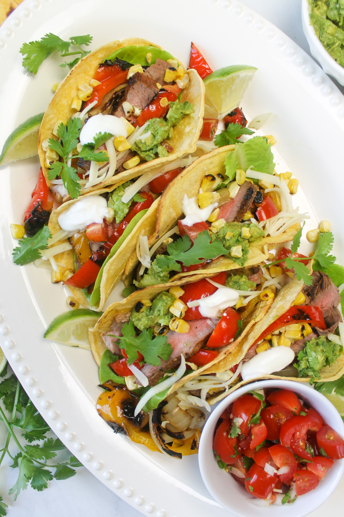 Grilled steak fajita tacos on a platter with a bowl of pico de gallo and garnished with cilantro and lime wedges.