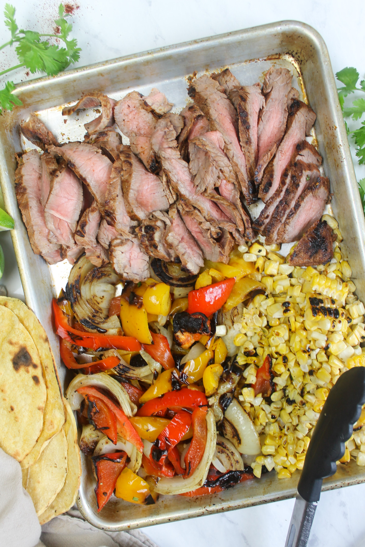 A sheet pan of sliced steak, peppers, onions, and charred corn cut off the cob.