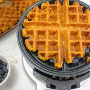Pumpkin waffle in an opened waffle iron with a sheet pan of waffles behind it.