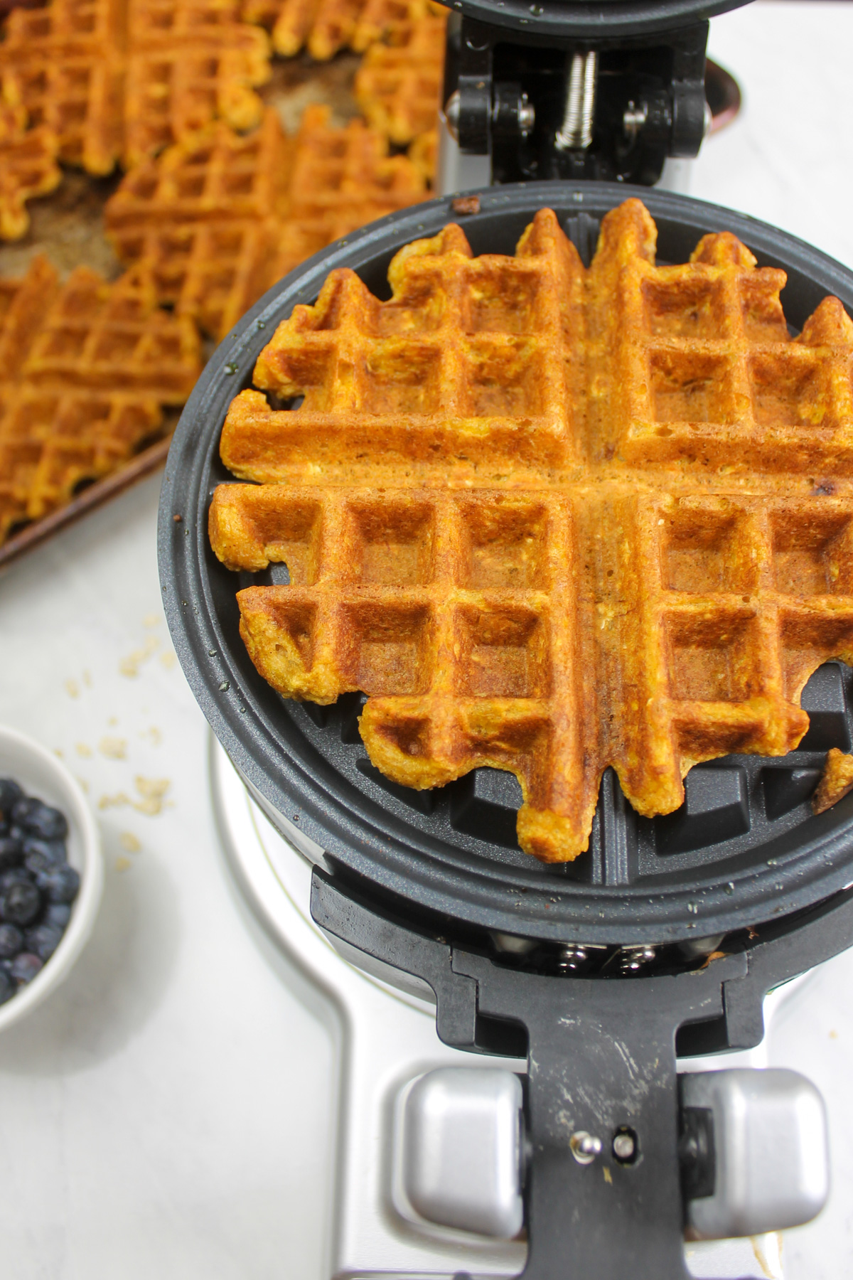 Pumpkin waffle in an opened waffle iron with a sheet pan of waffles behind it.