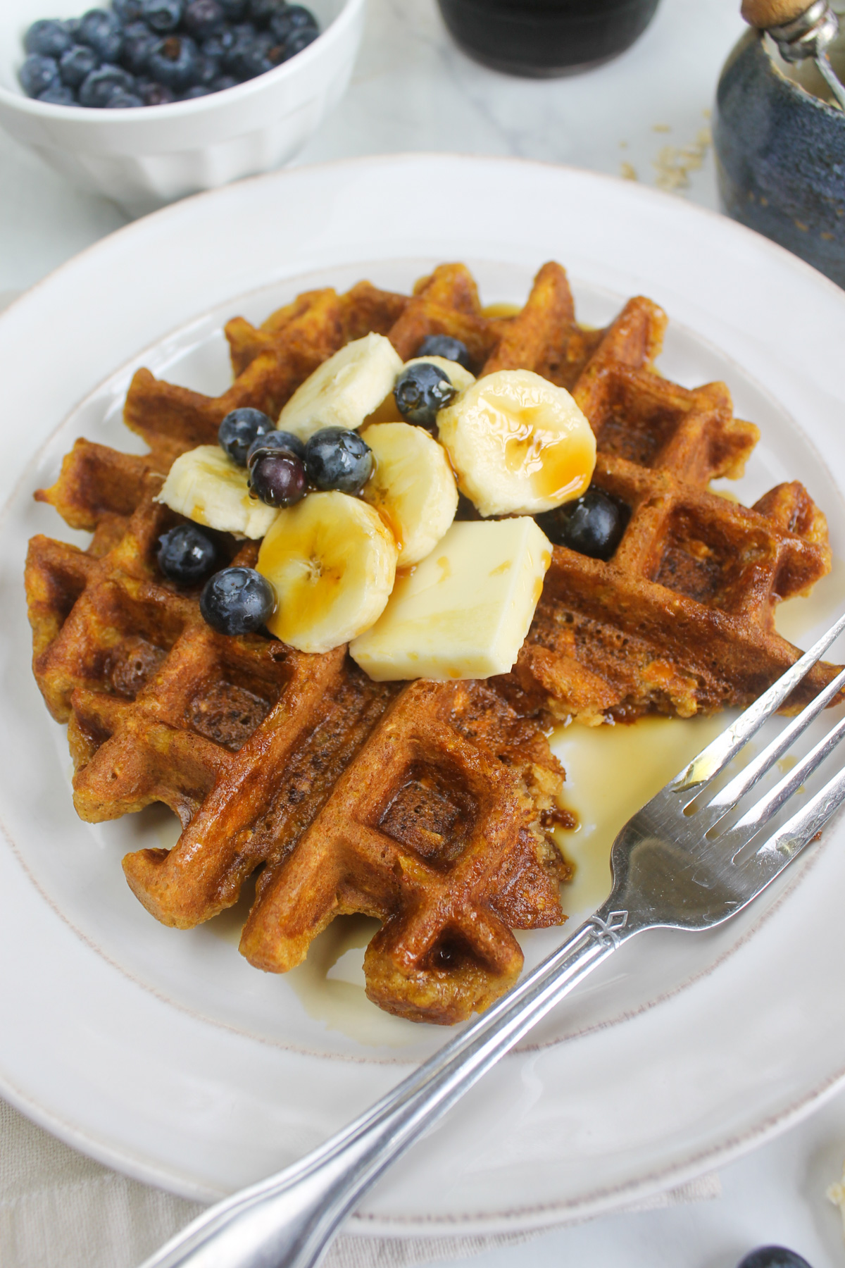 A pumpkin protein waffle with butter, maple syrup and topped with sliced bananas and blueberries.