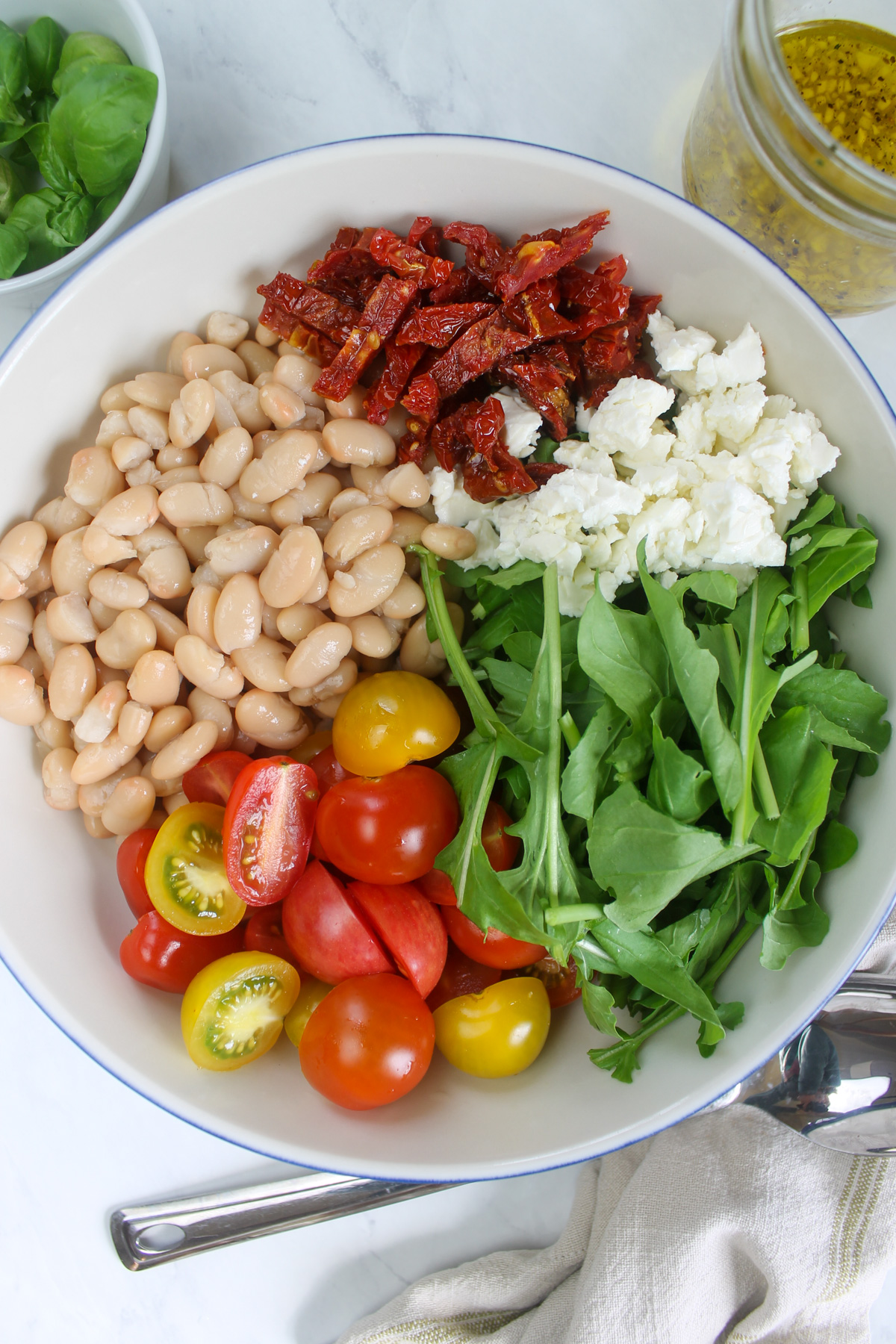 Adding all ingredients to a bowl including white beans, cherry tomato, sun-dried tomato, feta cheese, and arugula.