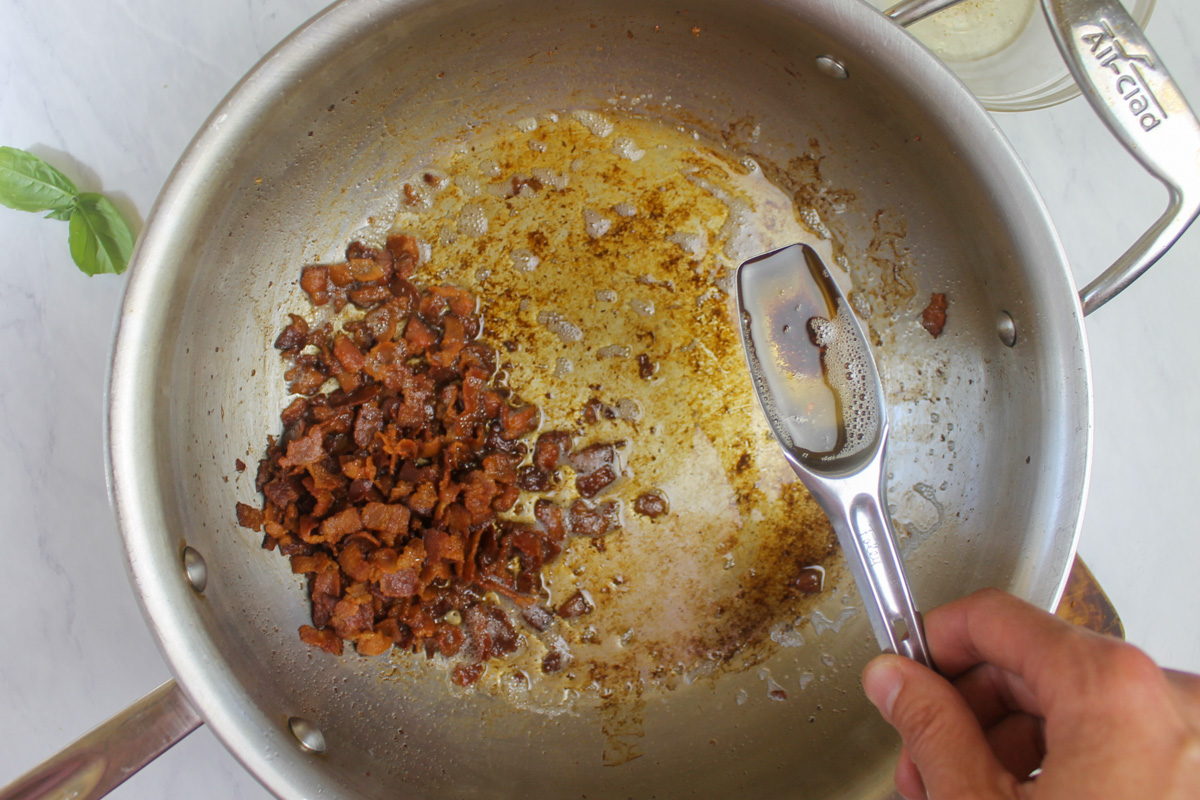 Chopped cooked crispy bacon, with a measuring spoon removing some of the bacon fat.