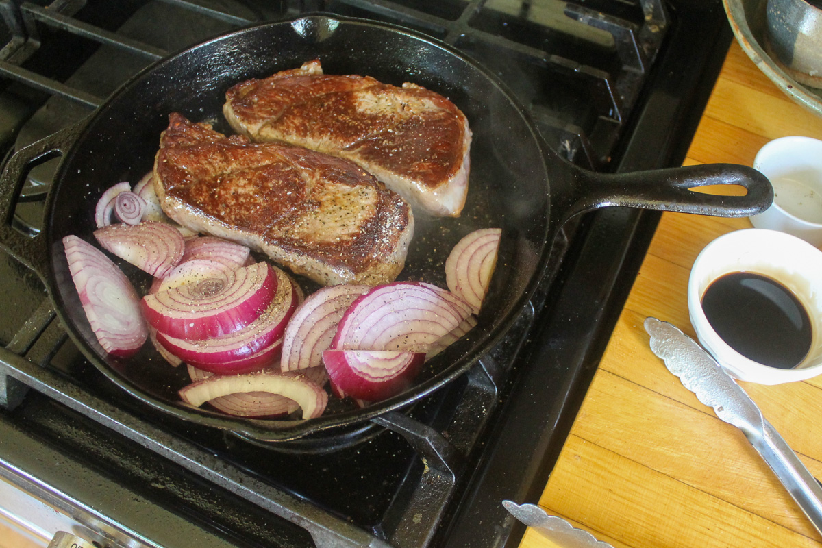 Steaks flipped in a cast iron skillet to sear the second side, raw red onion slices added to the pan next to the steak.