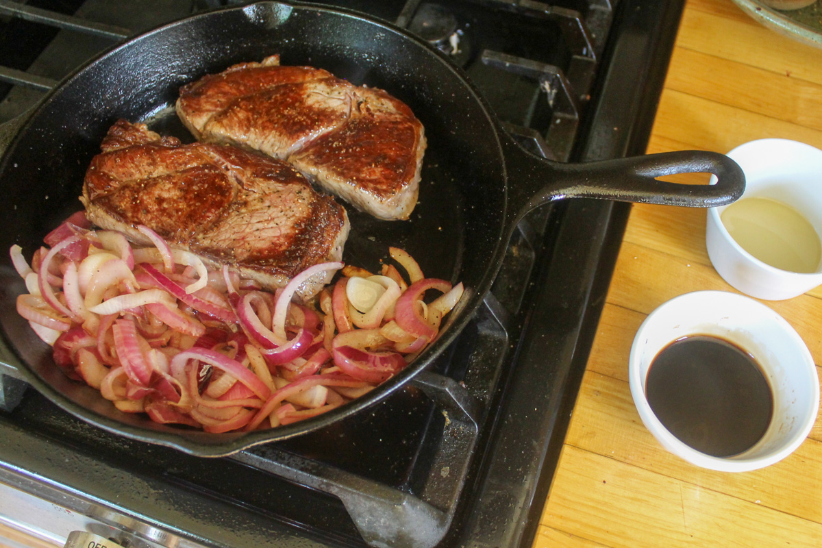 Steaks finishing cooking in a cast iron pan with onions caramelizing.