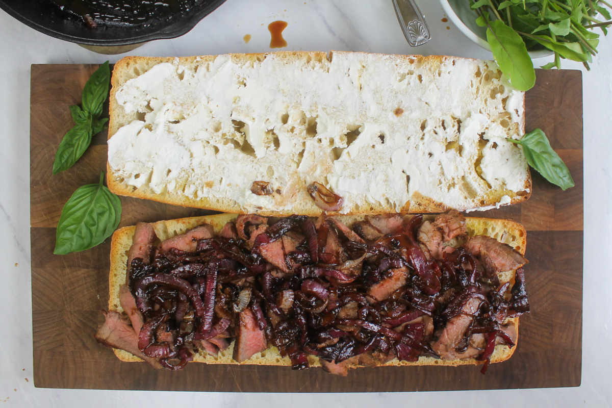 Assembling the sandwich on toasted ciabatta bread spread with coat cheese and piled with steak and balsamic onions.