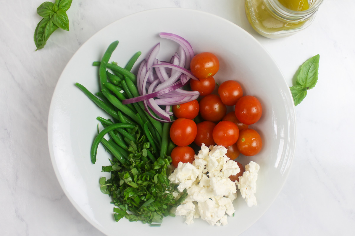 A white bowl with the green bean salad ingredients, including cherry tomato, feta cheese, red onion and herbs.