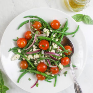 Green bean salad with cherry tomatoes, feta, red onion, and a jar of homemade vinaigrette dressing.