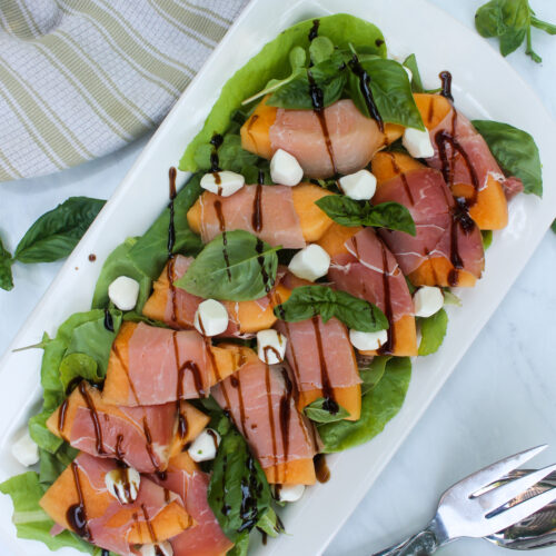 Melon wrapped in prosciutto on a platter of salad greens with mozzarella.
