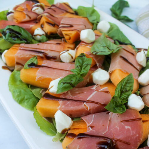 Melon wrapped in prosciutto on a platter of salad greens with mozzarella.
