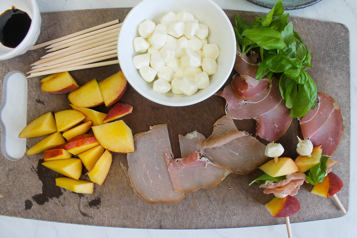 Ingredients prepared on a cutting board, peaches, mozzarella, prosciutto, basil and skewers.