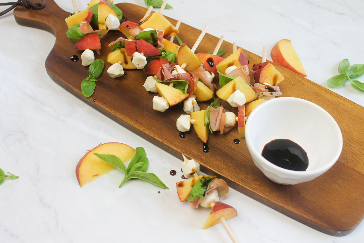 A wooden board of easy peach appetizer skewers with a small white bowl of extra balsamic glaze.