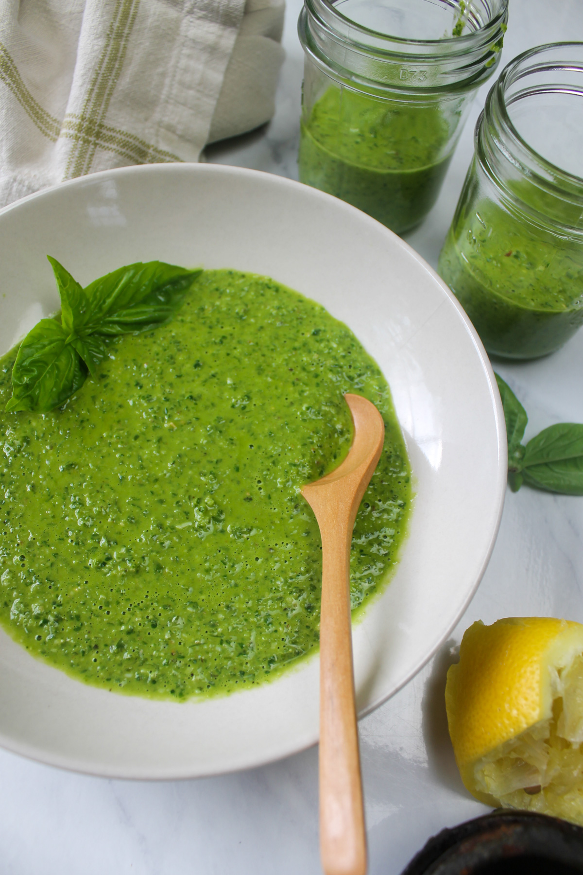 A white bowl of pesto with a wooden spoon with extra pesto in jars.