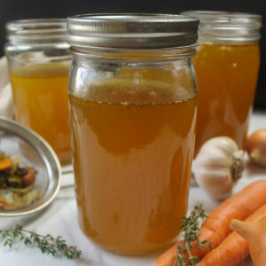 Quart jars of homemade chicken stock surrounded by carrots, thyme and garlic.