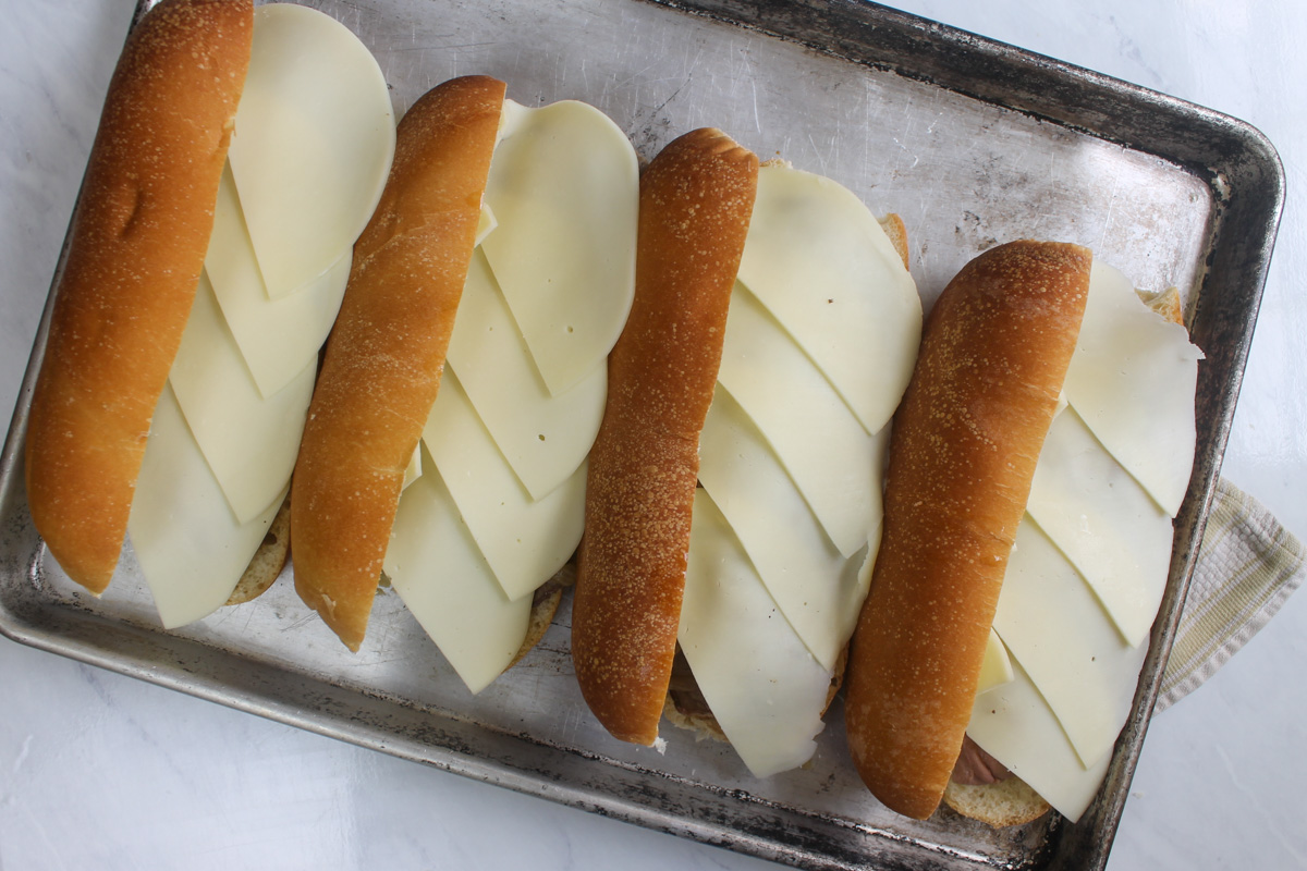 Topping French dip sandwiches with provolone cheese on a sheet pan.