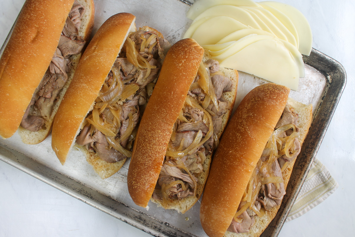 Assembling French dip sandwiches on hoagie buns on a sheet tray with roast beef and onions.