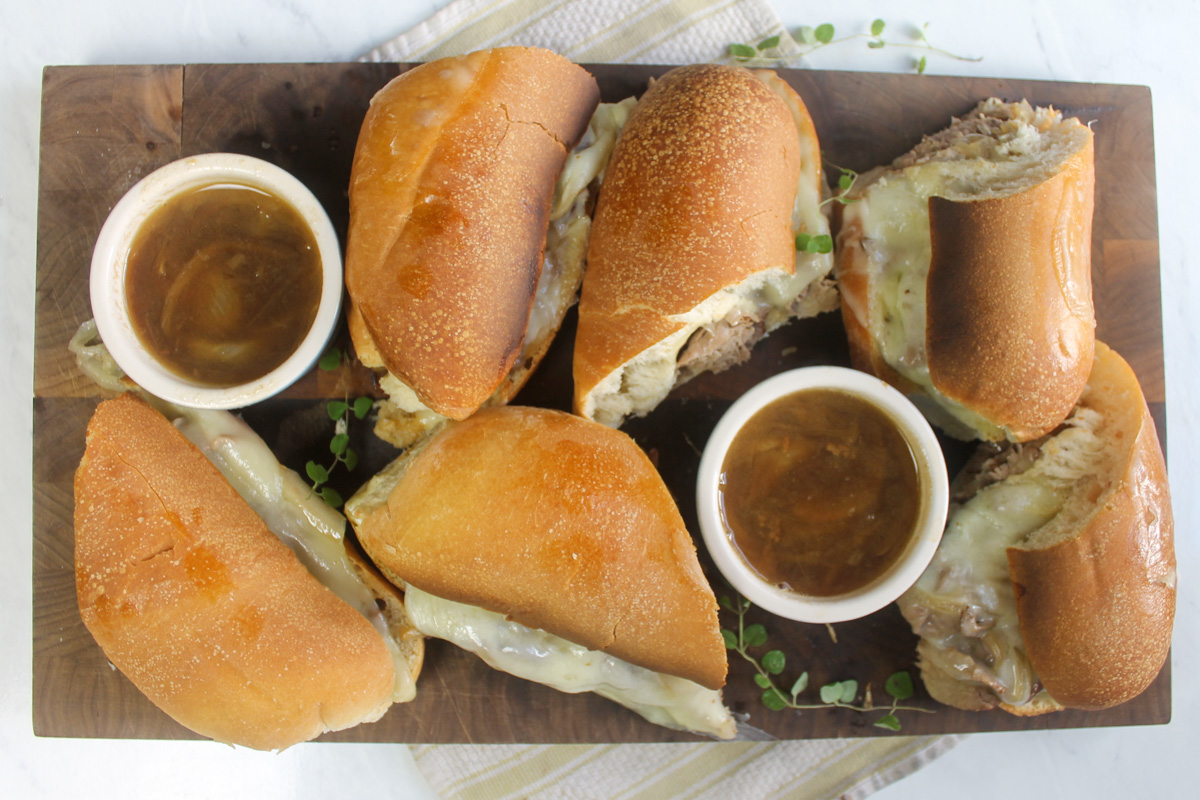 A cutting board platter of French dip sandwiches with cups of au jus dipping sauce.