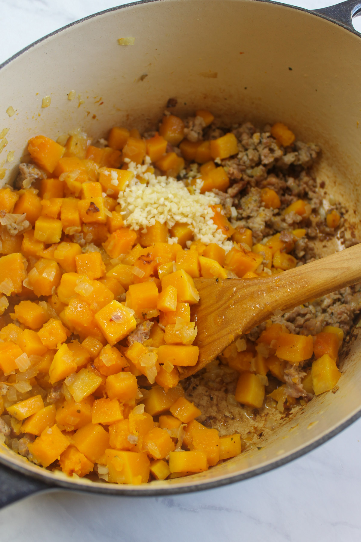A soup pot with browned sausage and sautéed butternut squash and onions, adding garlic.