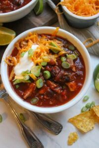 A white bowl of tomato based turkey chili with toppings and tortilla chips.
