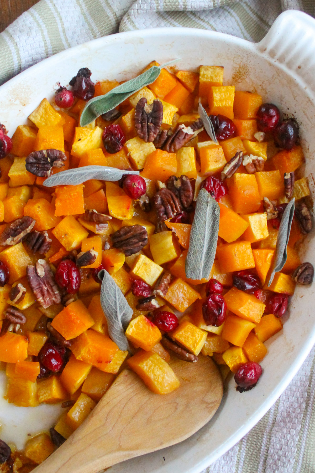 A white dish of maple roasted butternut squash with fresh cranberries and sage leaves.