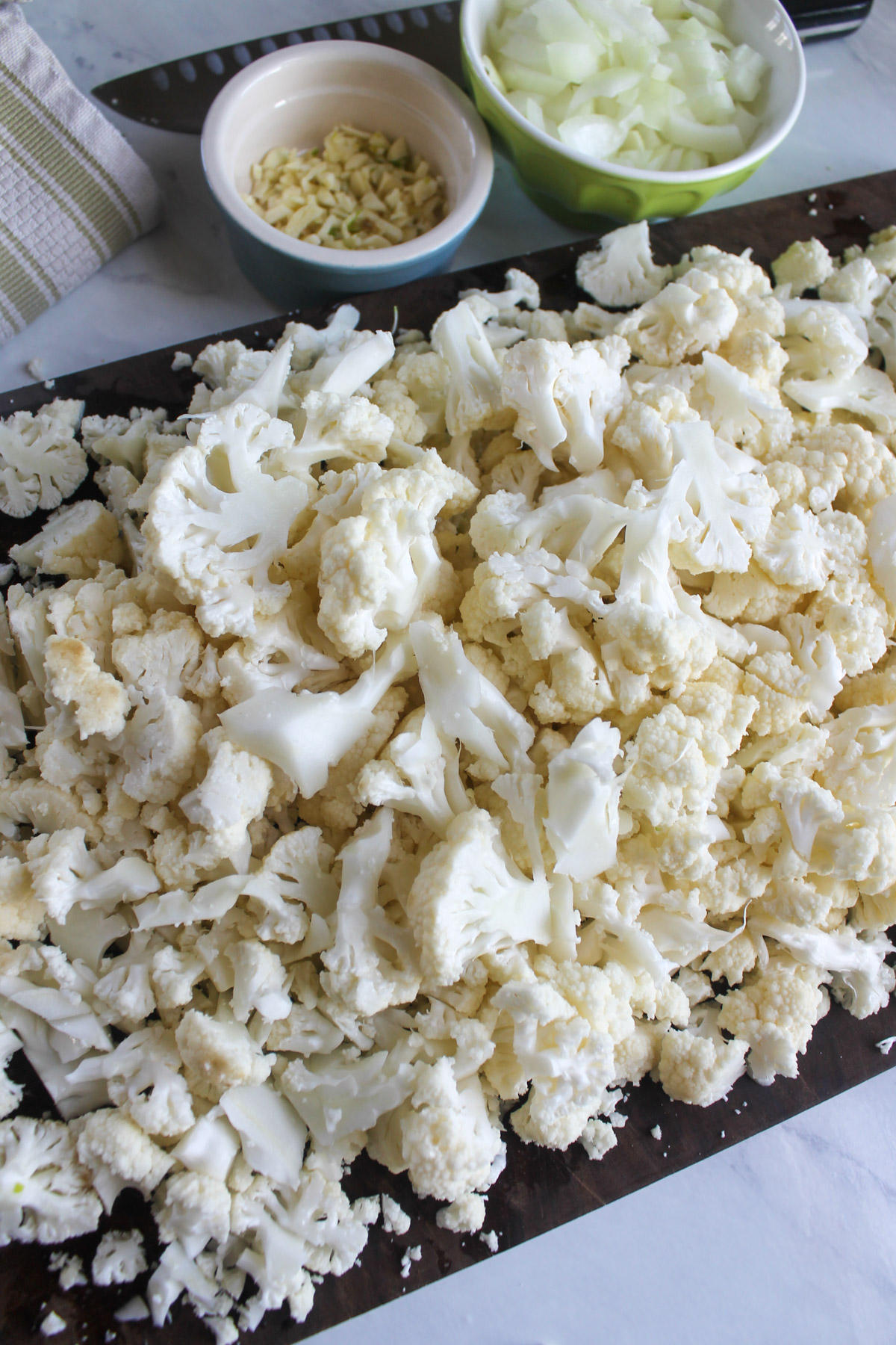 Chopped cauliflower on a cutting board with bowls of chopped onion and garlic.