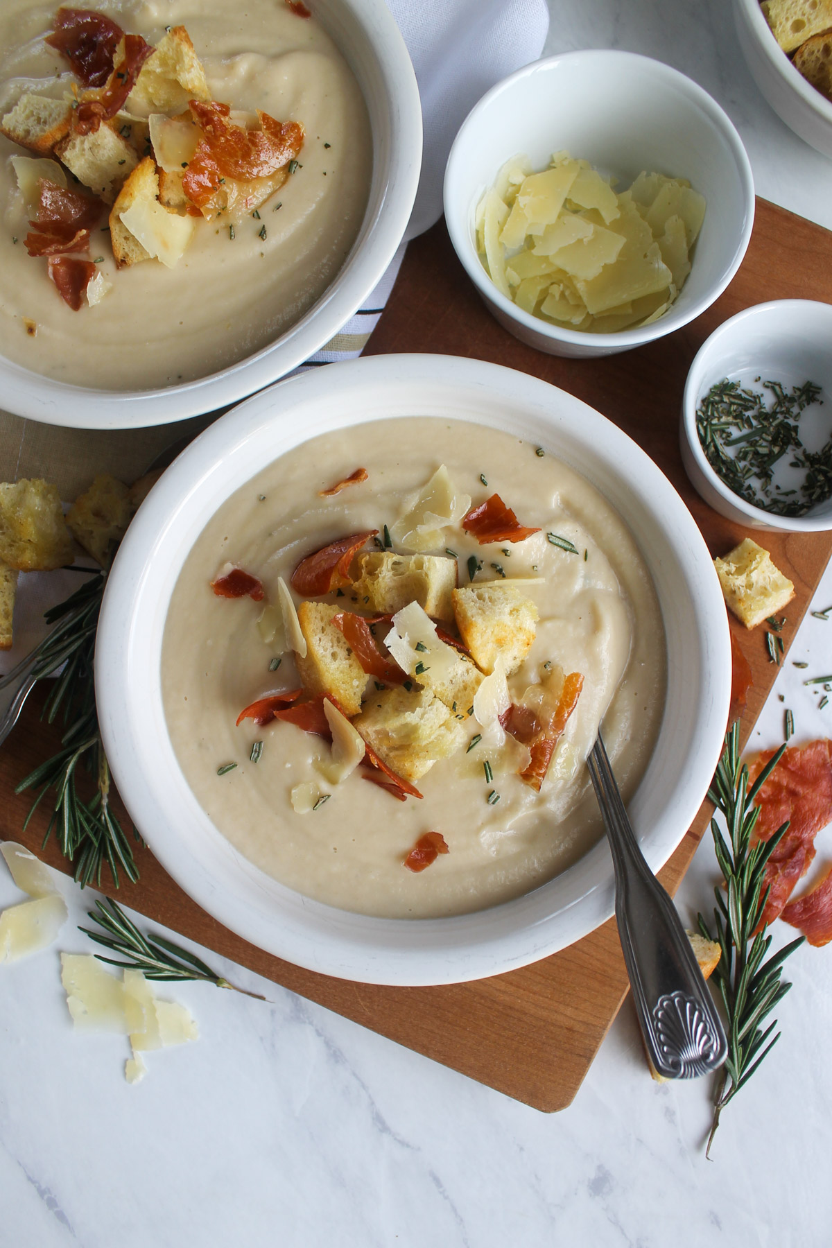 Two white bowls of cauliflower bean soup garnished with crispy prosciutto and homemade croutons.