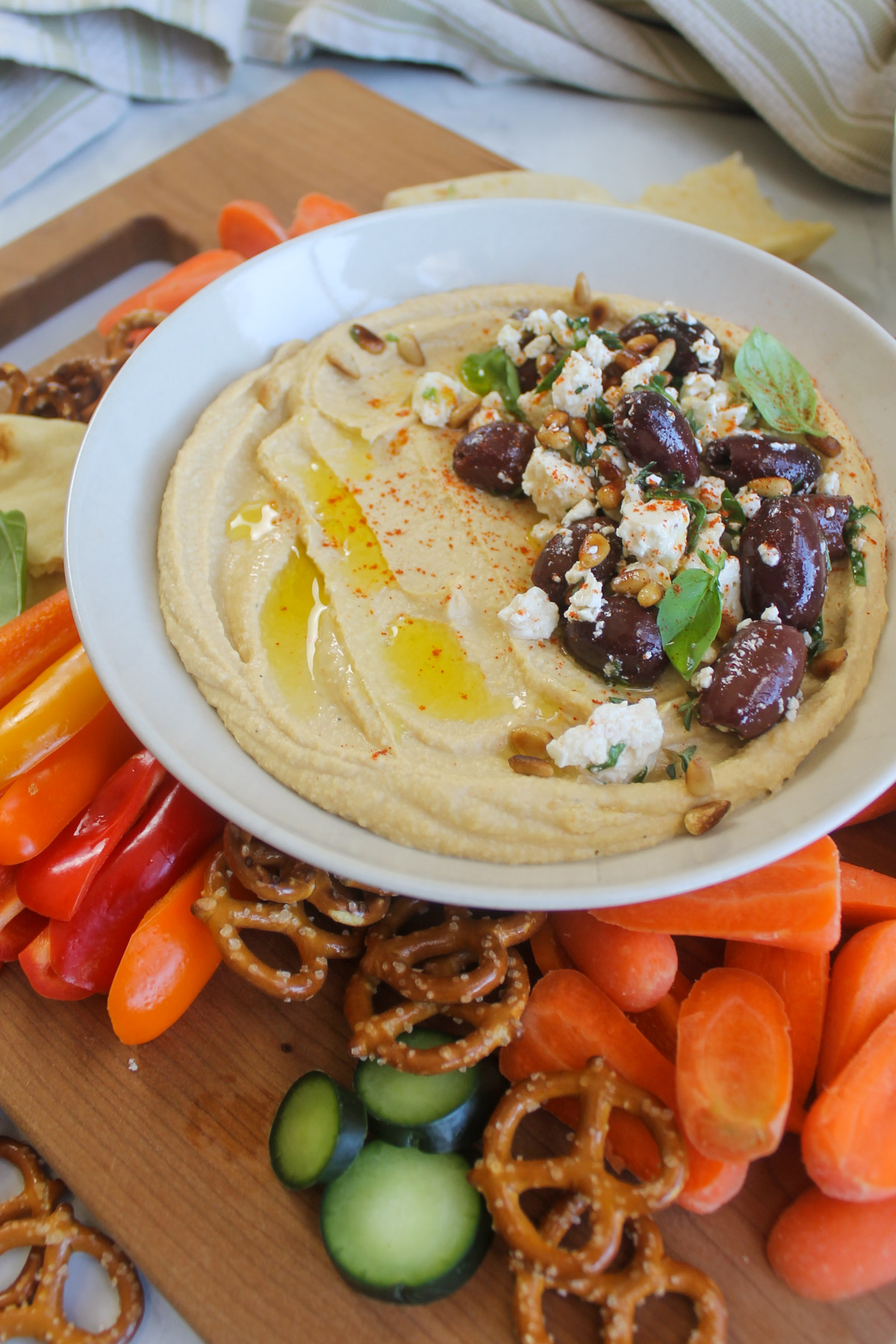 A white bowl of hummus with a topping made of pine nuts, olives and feta cheese on a board of veggies.