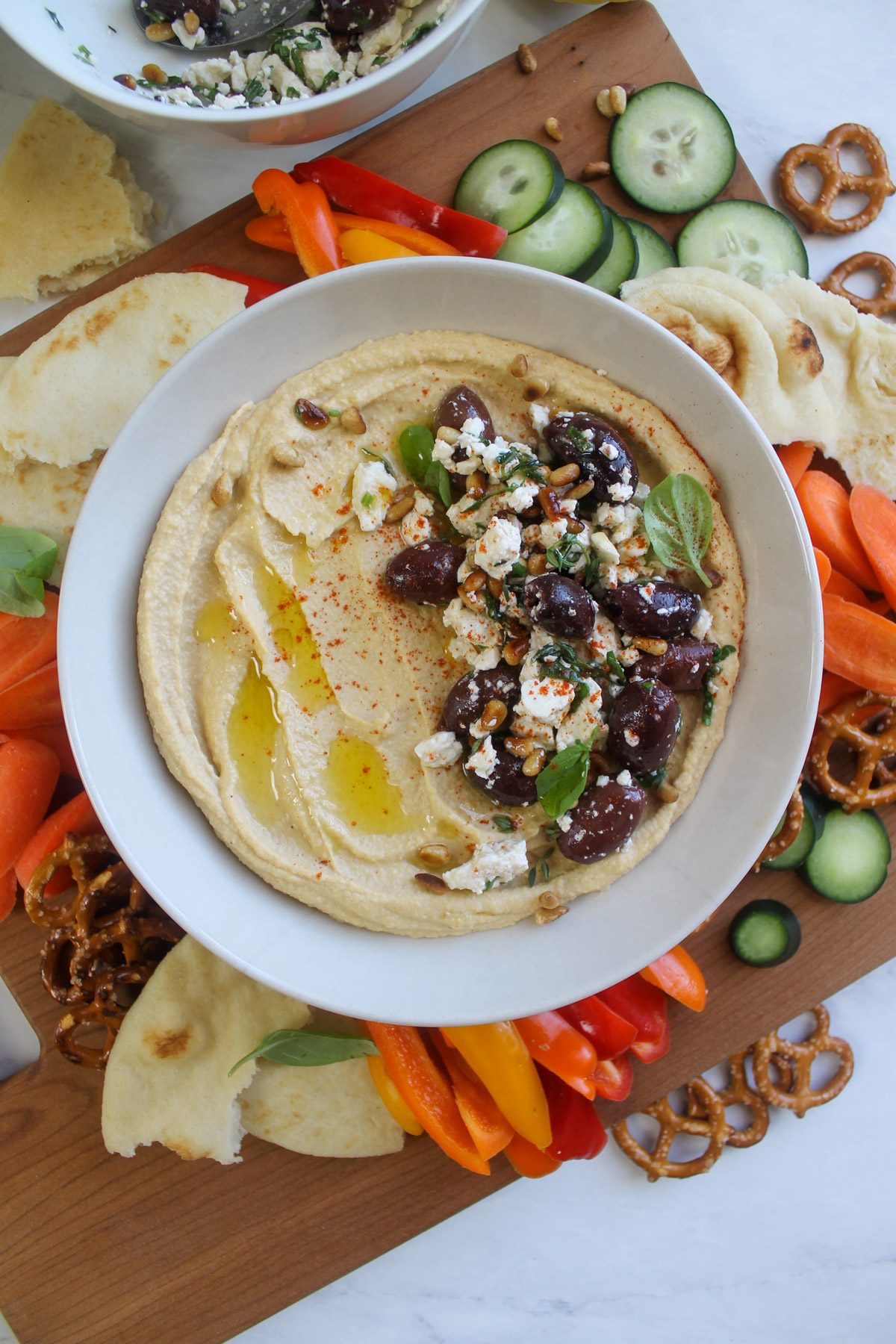 Homemade hummus in a bowl with pita, carrots, bell pepper, cucumbers, and pretzels to dip.