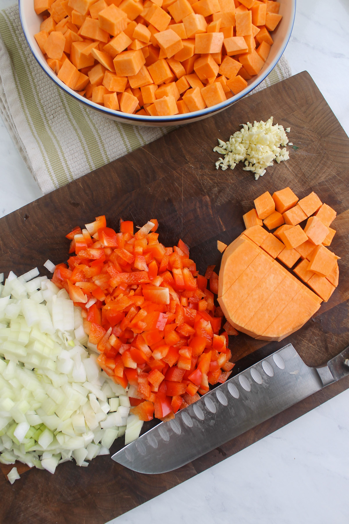Chopped sweet potato, red bell pepper, onion and garlic on a cutting board.