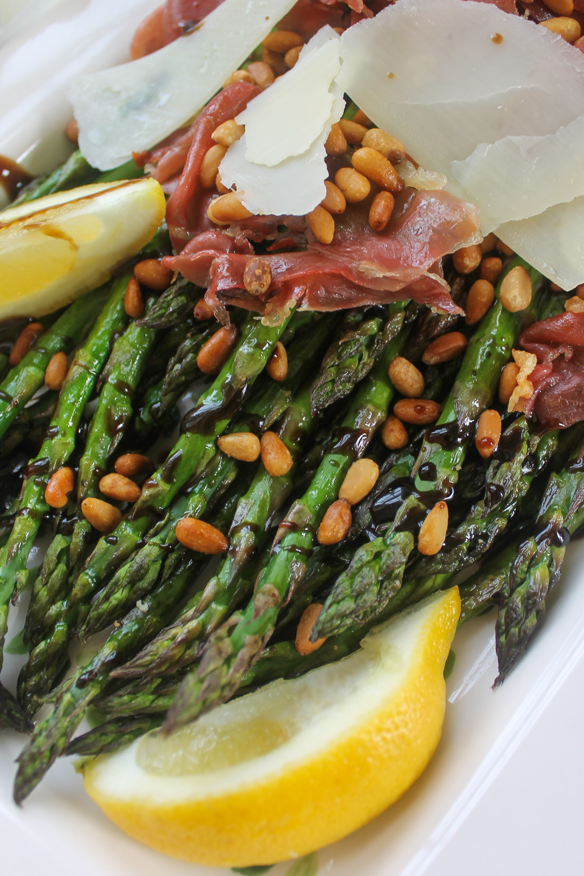 A close up of roasted asparagus tips with balsamic glaze and a topping made of crispy prosciutto and pine nuts.