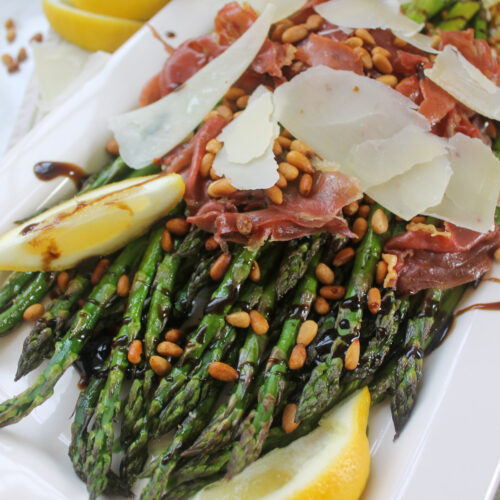 A platter of oven roasted asparagus spears topped with balsamic glaze and crispy prosciutto with pine nuts.