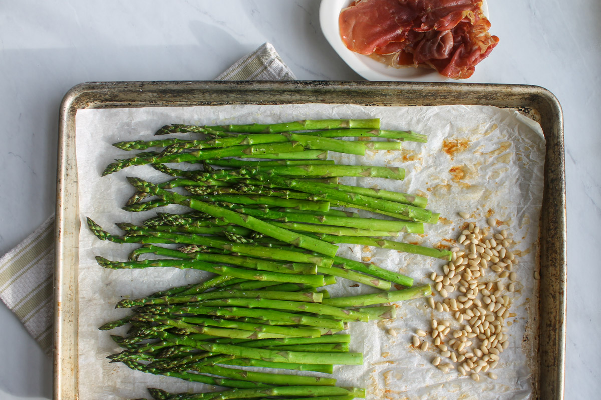 A sheet pan half way through roasting asparagus with the crispy prosciutto removed and pine nuts added.