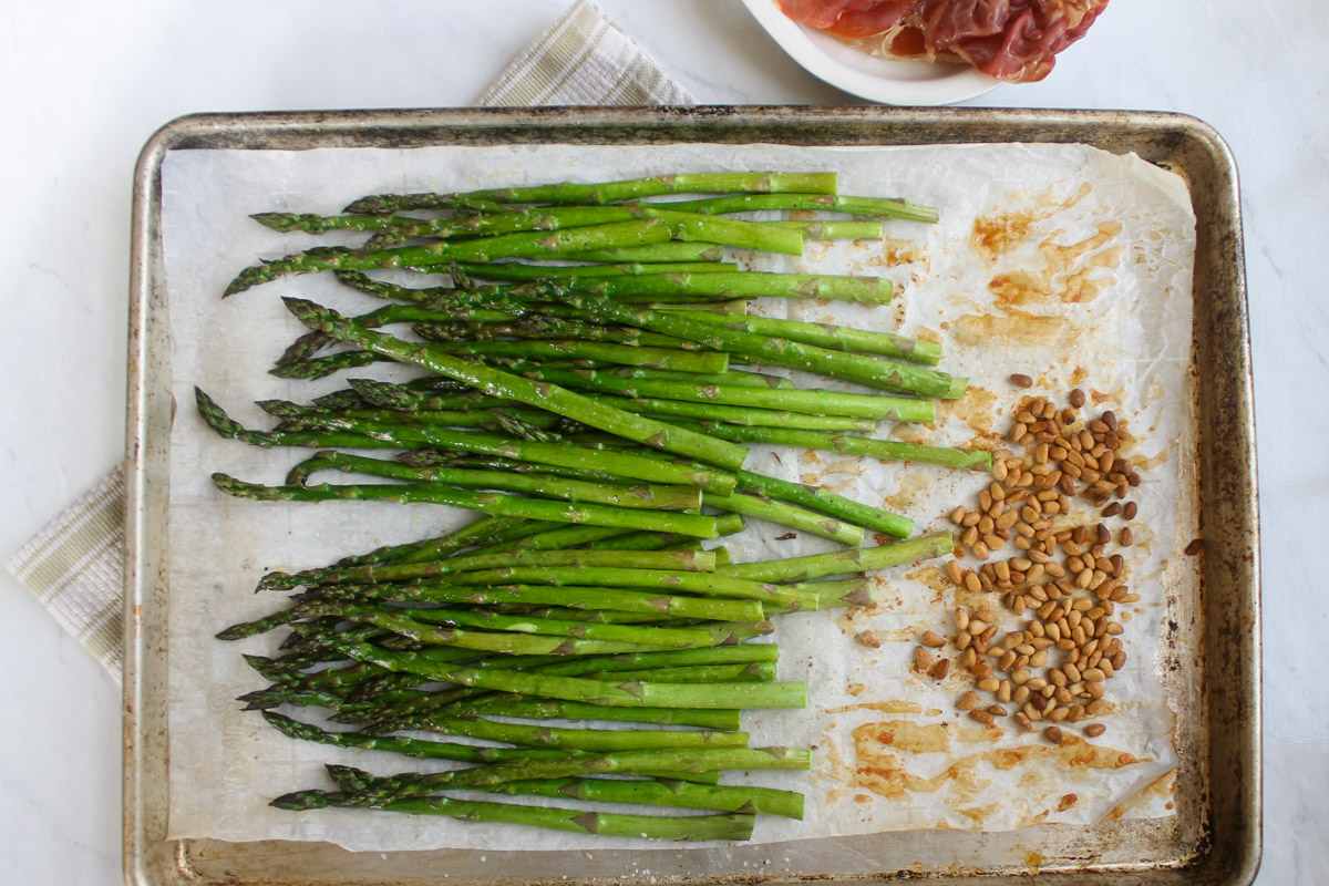 A sheet pan with roasted asparagus and toasted pine nuts with a bowl of crispy prosciutto.