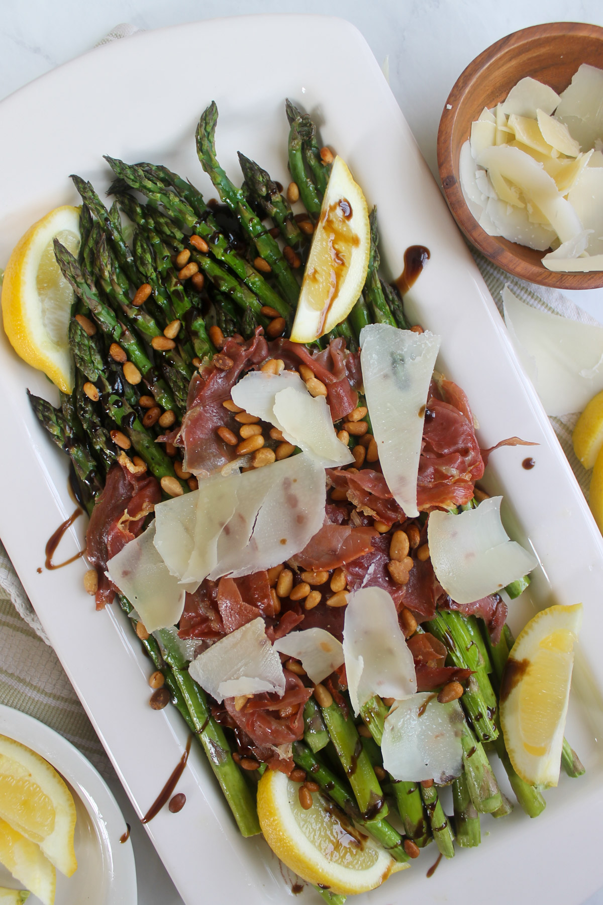 A white platter of roasted asparagus topped int he middle with crispy prosciutto, pine nuts and Parmesan cheese.