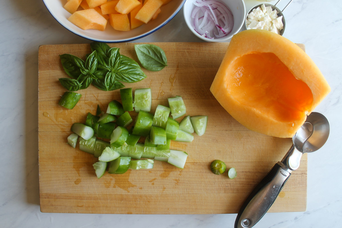 Chopped cucumber on a cutting board with a peeled and seeded half cantaloupe.