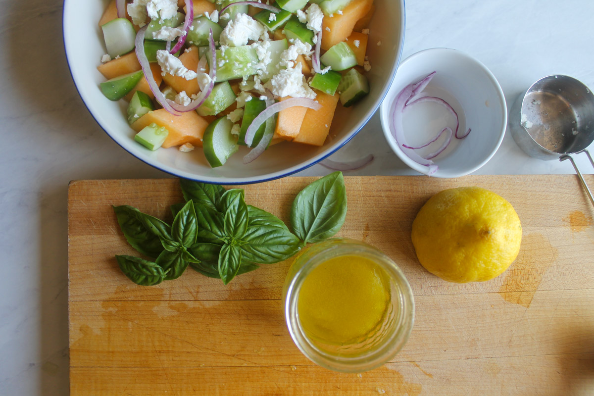 A jar of lemon vinaigrette on a cutting board with a bowl of cucumber melon salad.