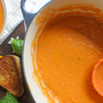 A soup pot of bright orange roasted tomato soup next to a bowl of soup and grilled cheese.