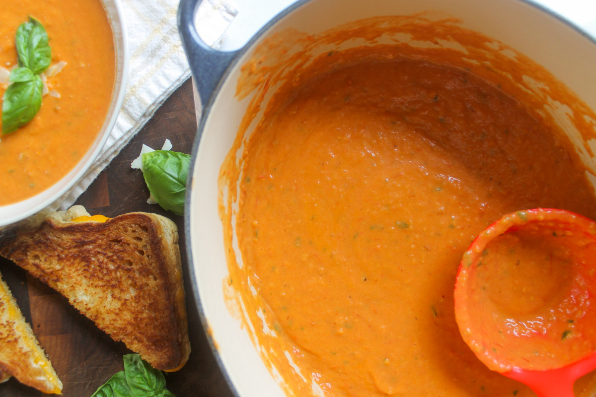 A soup pot of bright orange roasted tomato soup next to a bowl of soup and grilled cheese.