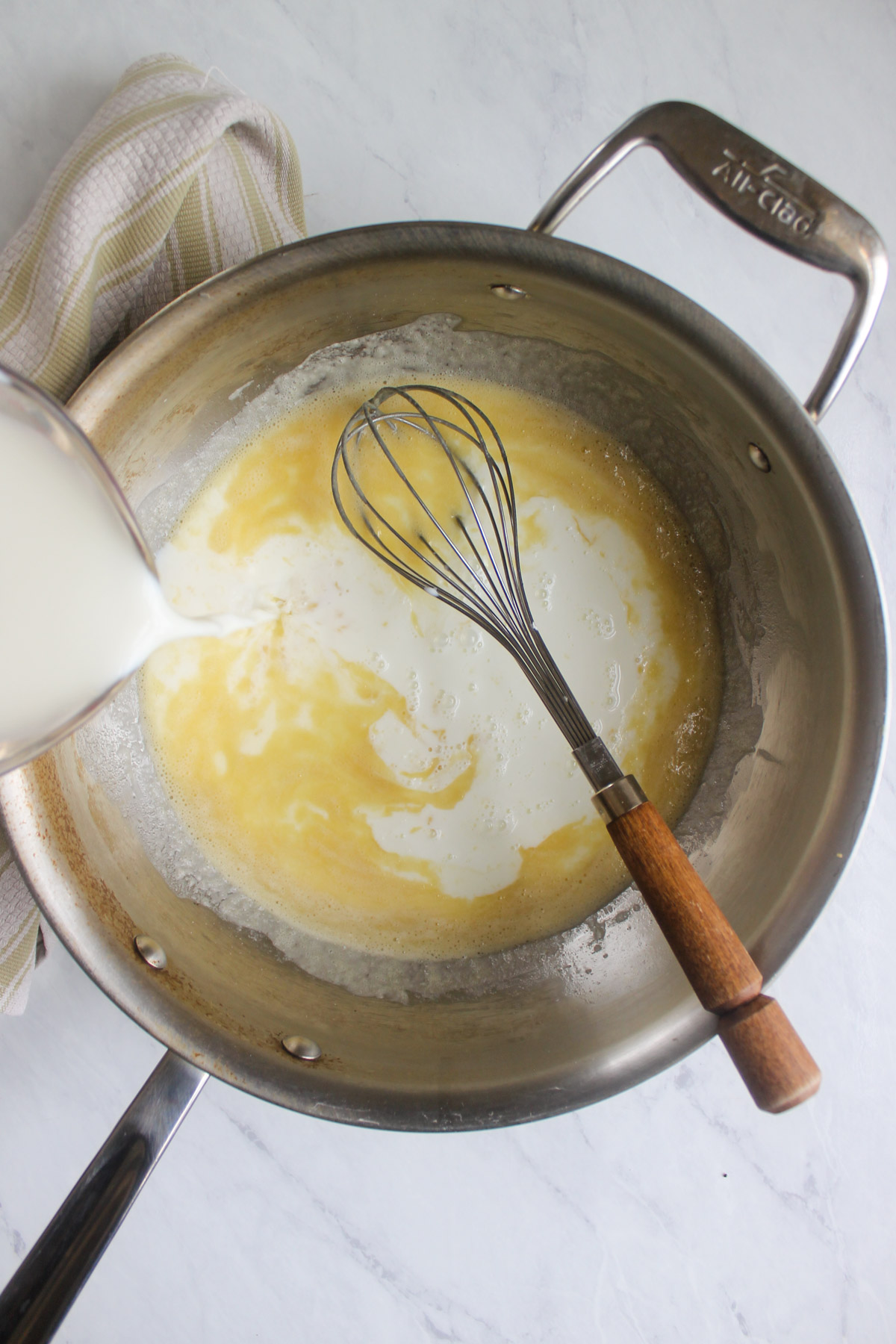 Adding milk to a roux of butter and flour in a skillet with a whisk.